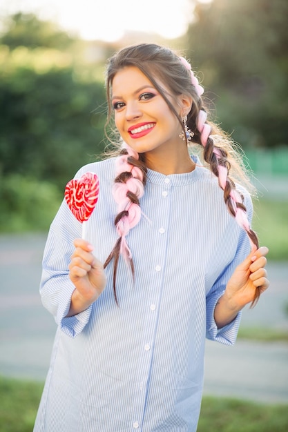 Pretty girl with colorful braids in blue dress, holding candy heart on stick.