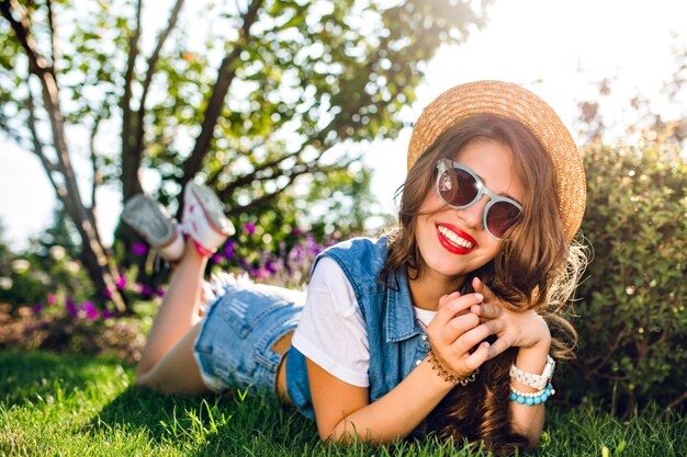 Pretty girl with long curly hair in hat is lying on grass in summer park. on sunlight. She wears jeans jerkin, shorts, sunglasses. She is smiling to camera with red lips.