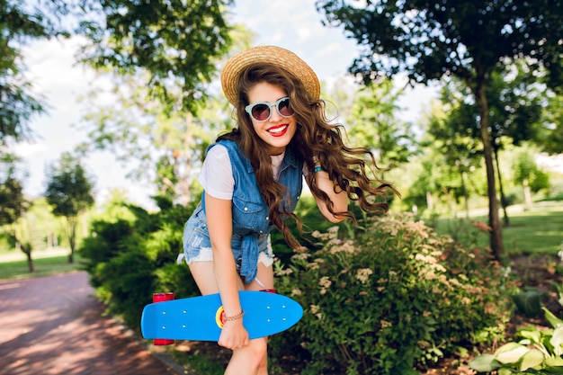 Pretty girl with long curly hair and red lips is posing with skateboard in summer park. She wears jeans jerkin, sunglasses, hat.