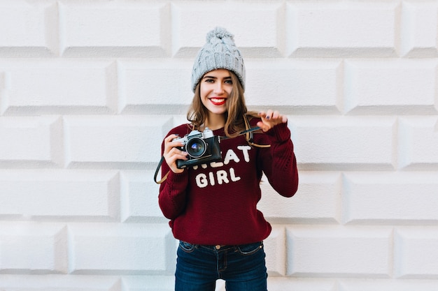 Free photo pretty girl with long hair on grey wall . she wears knitted hat, holds camera and smiling friendly.