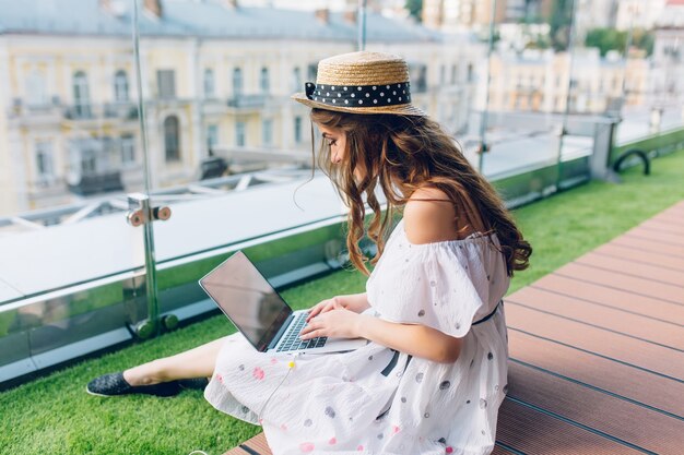 Pretty girl with long hair is sitting on floor on the terrace. She wears a white dress with naked shoulders and hat . She is typing on  laptop on knees.