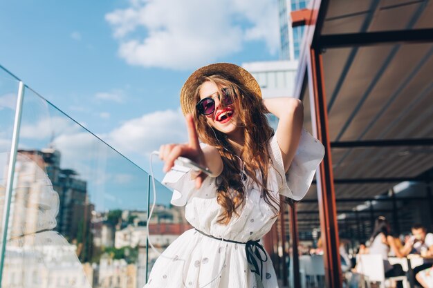 Pretty girl with long hair  in sunglasses is listening to music  on the terrace. She wears a white dress, red lipstick and hat . She is stretching hand to the camera and dancing. Buttom view.