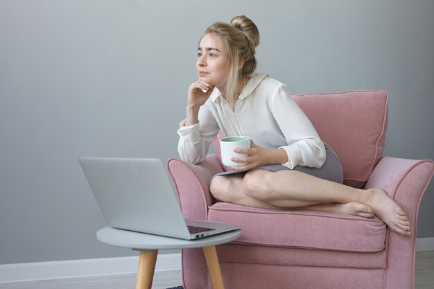 Pretty girl with messy hairstyle having dreamy facial expression, enjoying coffee, sitting in comfortable armchair in front of open portable computer, thinking about future plans and summer vacations