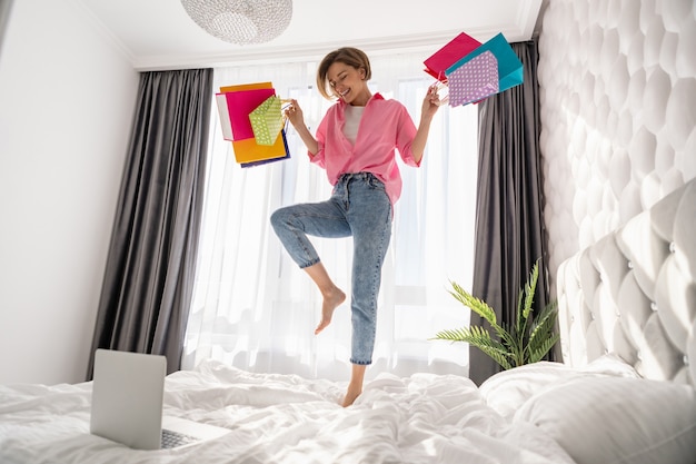 Free photo pretty happy woman having fun jumping on bed at home with colorful shopping bags