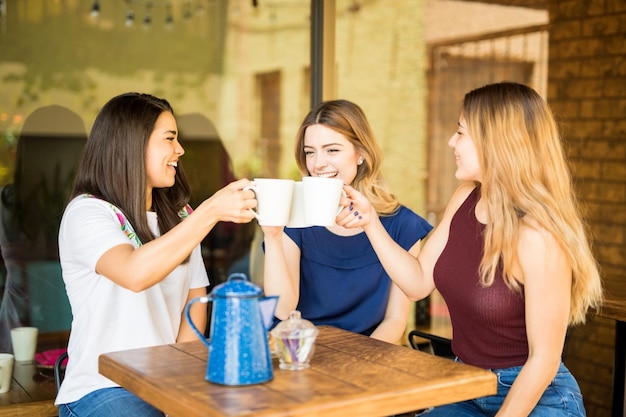 Pretty hispanic female friends having fun and making a toast with coffee cups at restaurant