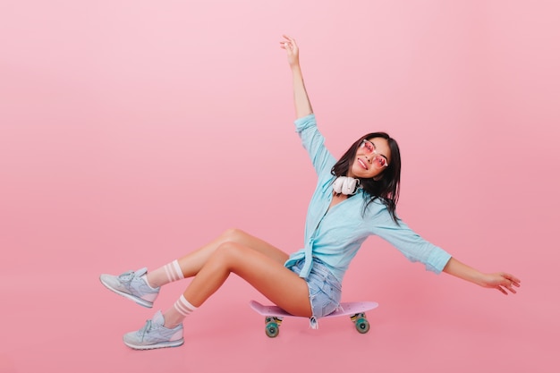 Pretty hispanic woman with bronze skin waving hands while sitting on longboard. inspired latin girl in sunglasses posing on skateboard