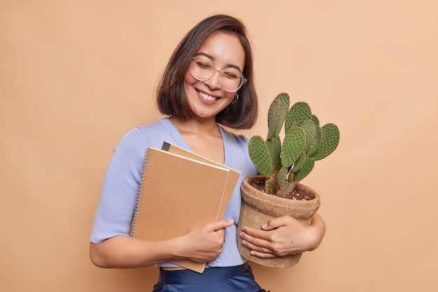 Pretty joyful Asian student feels satisfied after passing exam carries notebooks and potted cactus wears transparent glasses casual jumper poses against beige wall