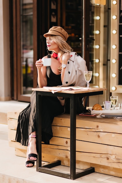 Free photo pretty lady in long dress and black sandals enjoying lunch in outdoor cafe and looking away. fascinating blonde girl in hat waiting friend to eat croissants together.