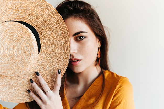 Free photo pretty lady mysteriously on white wall. portrait of model in yellow blouse with boater in hands.