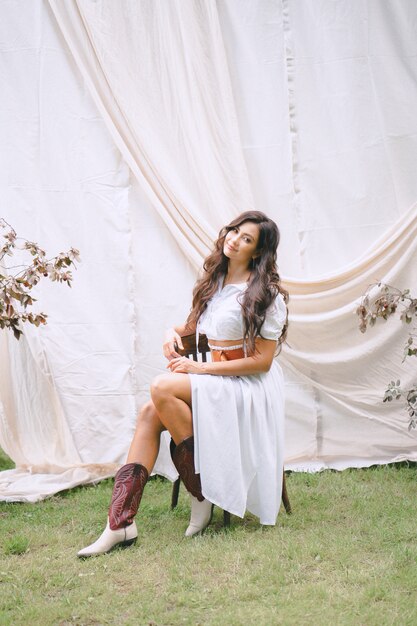 Pretty lady sitting on a chair and looking, in garden with white wall, in long white dress with belt during daytime .