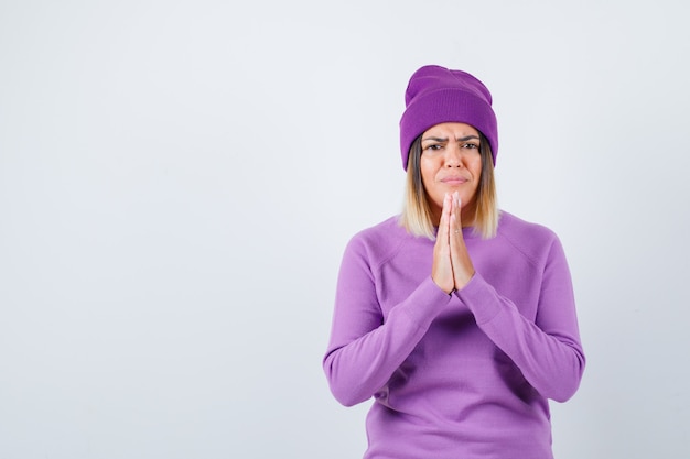 Pretty lady with hands in praying gesture in sweater, beanie and looking cheerless. front view.