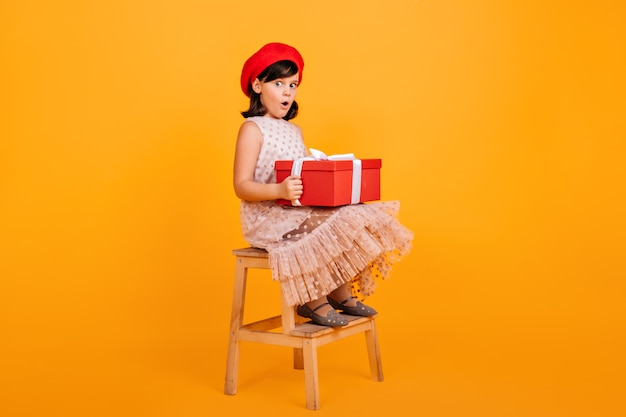 Pretty little girl in dress sitting on chair and holding big present box.  french kid with birthday gift.