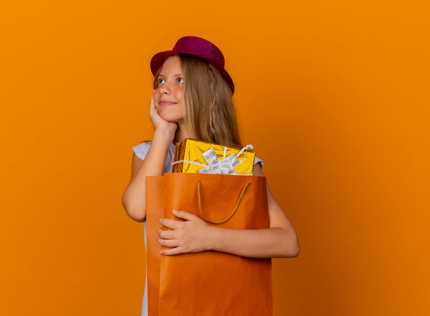 Pretty little girl in holiday hat holding paper bag with gifts looking aside with happy face, birthday party concept standing over orange background