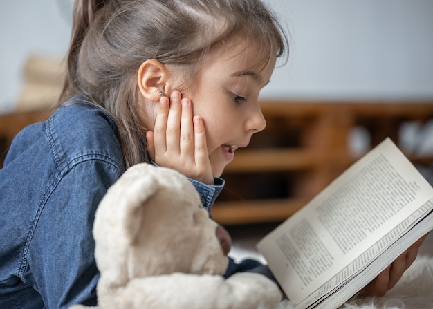 Pretty little girl at home, lying on the floor with her favorite toy and reads book.