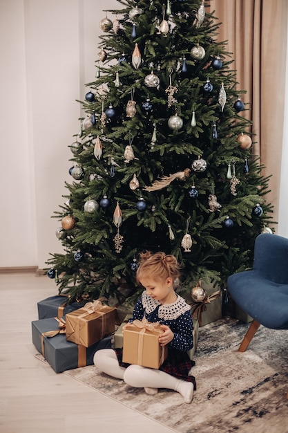 Pretty little girl sits under a big christmas tree, holds her present and smiles