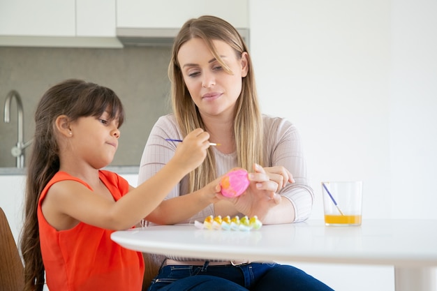 Pretty mom holding Easter egg and her daughter drawing on it.