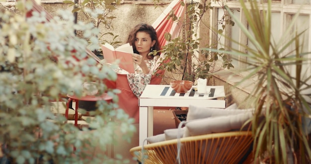 Free photo pretty redhaired young woman in floral dress reads a book in a hammock in a small urban patio garden