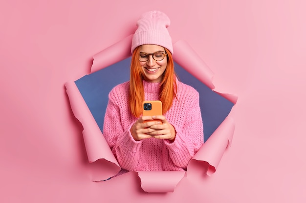 Pretty redhead woman looks happily at smartphone device checks her email smiles pleasantly dressed in stylish clothing.