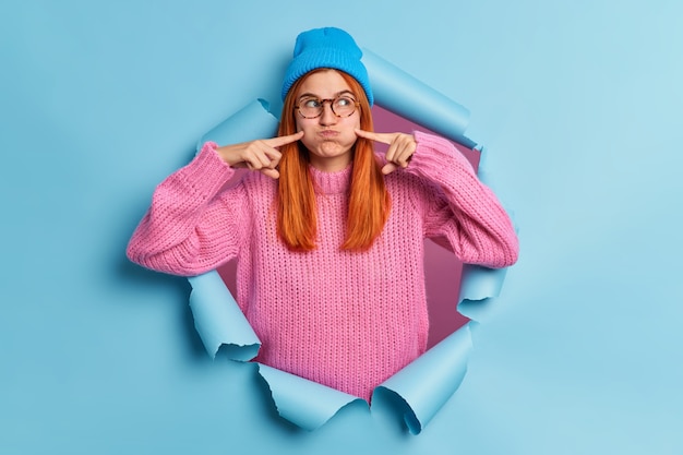 Pretty redhead woman makes funny grimace blows cheeks and points with index fingers holds breath wears blue hat knitted sweater.