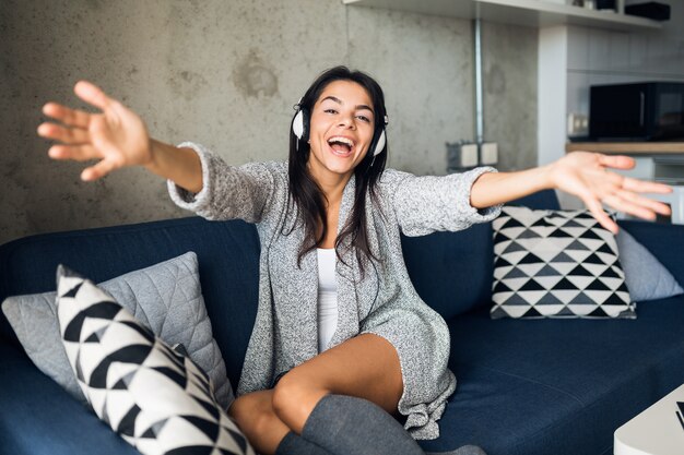 Pretty sexy smiling woman in casual outfit sitting in living room listening to music on headphones, having fun at home