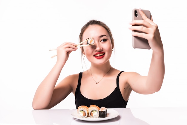 Pretty smiling asian woman cover her eye with sushi roll and make selfie on her phone