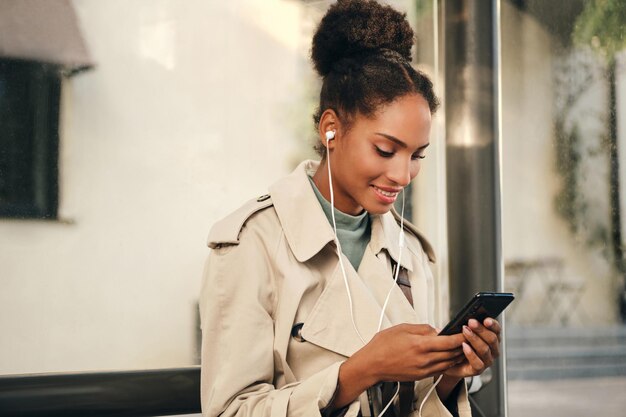 Pretty smiling casual African American girl in stylish trench coat and earphones happily using cellphone at bus stop