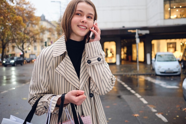 Pretty smiling girl happily talking on smartphone walking around cozy city street with shopping bags