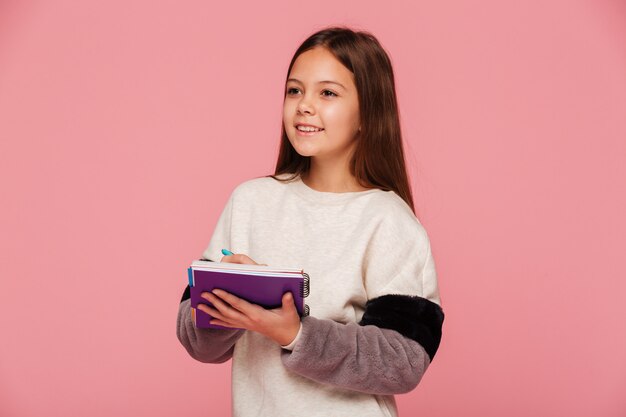 Pretty smiling girl looking aside and writing in notebook isolated