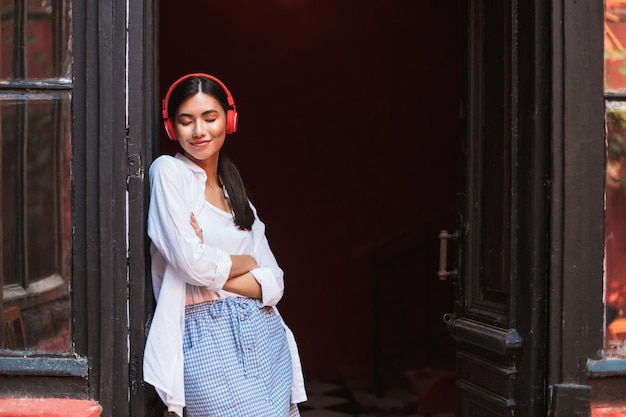 Free photo pretty smiling girl in white shirt happily closing eyes leaning on old door while listening music in red headphones