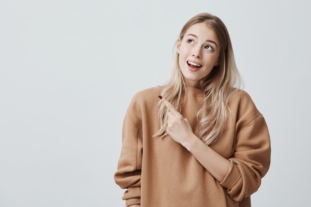 Pretty smiling joyfully female with fair hair, looking upwards, pointing index fingers, showing copy space