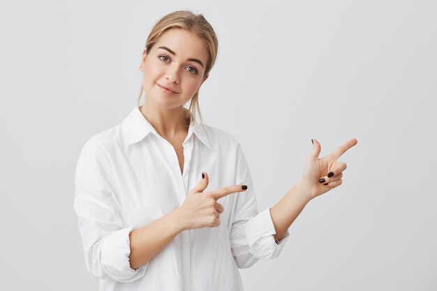 Pretty smiling joyfully female with fair hair, pointing her index fingers, showing copy space for advertising content. Studio shot of good-looking beautiful girl isolated on studio wall.