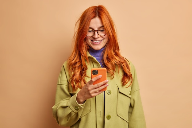 Free photo pretty smiling redhead woman uses mobile phone application glad to get message from boyfriend has pleasant conversation online dressed in fasionable autumn clothes.