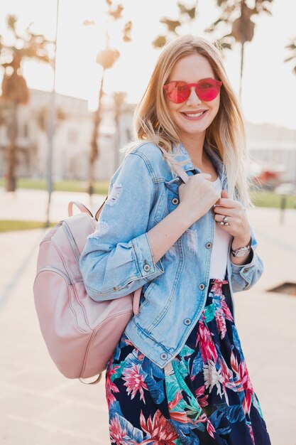 Pretty smiling woman walking in city street in stylish printed skirt and denim oversize jacket wearing pink sunglasses, holding leather backpack, summer style trend