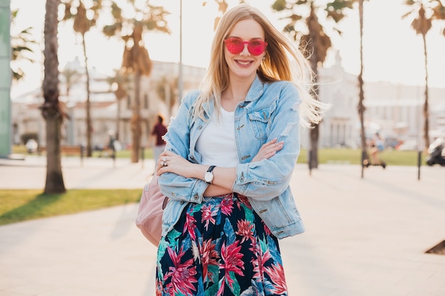 Pretty smiling woman walking in city street in stylish printed skirt and denim oversize jacket wearing pink sunglasses, summer style trend