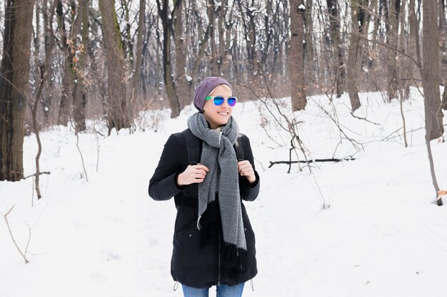 Pretty smiling woman with warm clothing holding backpack standing in snowy landscape during winter