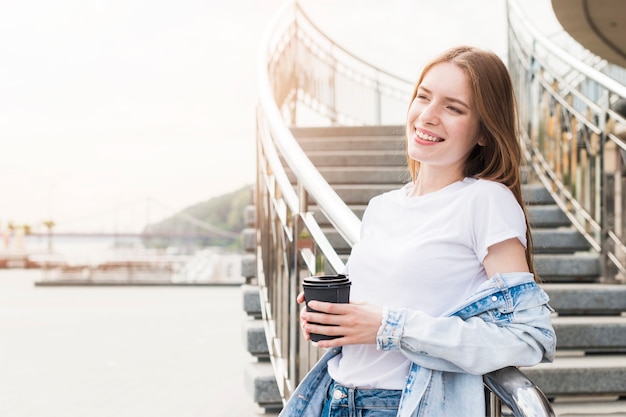 Pretty smiling young woman leaning on metallic railing with holding disposable cup