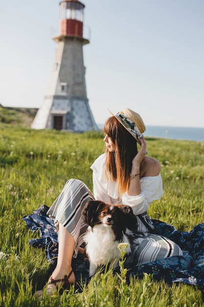 Pretty stylish woman in countryside, holding a dog, happy positive mood, summer, straw hat, bohemian style outfit