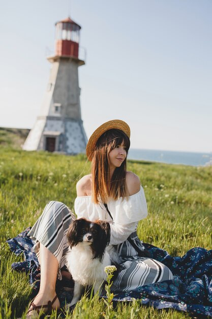 Pretty stylish woman in countryside, holding a dog, happy positive mood, summer, straw hat, bohemian style outfit