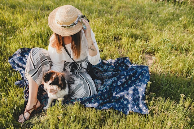 Pretty stylish woman in countryside, holding a dog, happy positive mood, summer, straw hat, bohemian style outfit