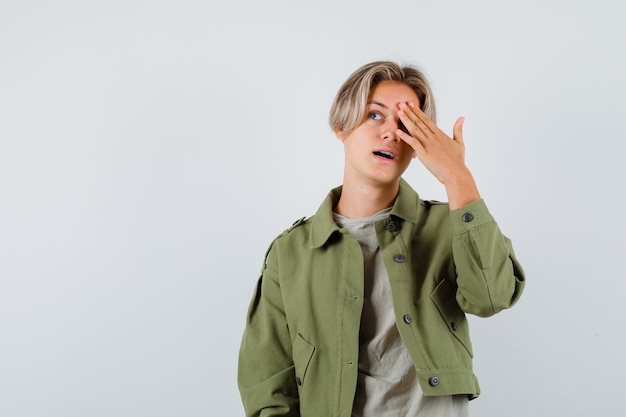 Pretty teen boy keeping hand on eye, looking up in green jacket and looking wondered , front view.