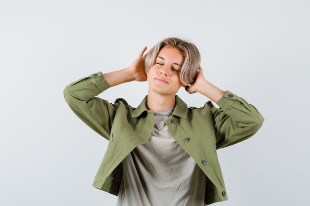 Free photo pretty teen boy keeping hands behind ears, shutting eyes in green jacket and looking delighted , front view.