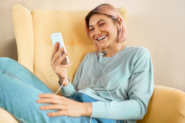 Pretty teen girl relaxing at home chatting with friends online on mobile via group webcam call, smiling. Cute young woman sitting in armchair with cell phone and earbuds, watching funny video