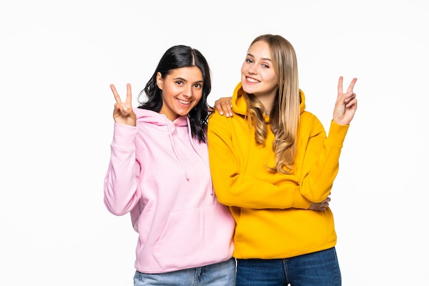 Free Photo pretty two girlfriends women showing v-sign symbols, wearing casual bright hoodies and jeans isolated white wall