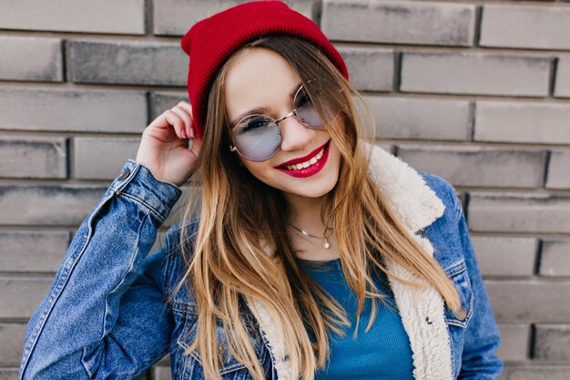 Pretty white girl with cheerful smile having fun in cold spring day. Outdoor portrait of joyful blonde woman wears blue glasses and red hat.