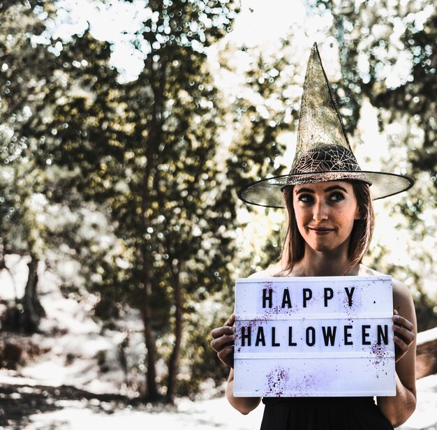 Pretty witch with greeting signboard in sunny forest