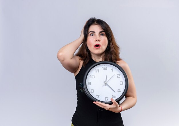 Pretty woman in black blouse holding clock lookin worried for being late 