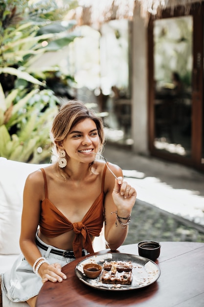 Free Photo pretty woman in brown cropped top bites plastic fork, smiles and enjoys delicious waffle with chocolate syrup