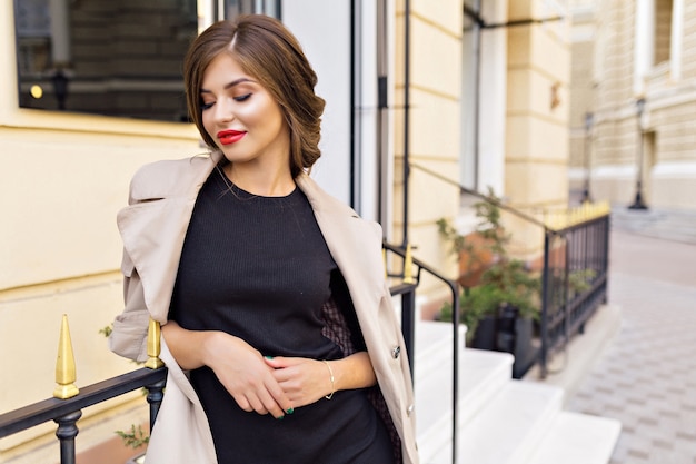 Free photo pretty woman dressed in black dress and beige trench with stylish hairstyle and red lips at the street