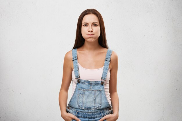 Pretty woman of European appearance dressed in denim jumpsuit holding her breath, trying hard not to laugh.