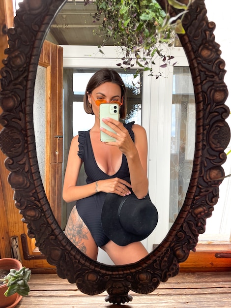 Free Photo pretty woman at home takes photo selfie in mirror on mobile phone for stories and posts in social media, wearing black summer swimsuit